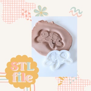 May include: A light brown clay mold with a turtle design pressed into it, next to a white turtle-shaped mold. The image also includes the text "STL file" in a yellow and orange design.
