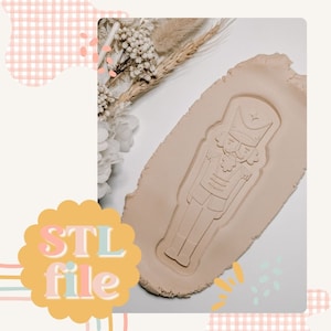 May include: A beige play dough with a nutcracker design pressed into it. The design is outlined in white and the nutcracker is wearing a hat with a star on top. The words "STL file" are written in colourful letters on a yellow circle.