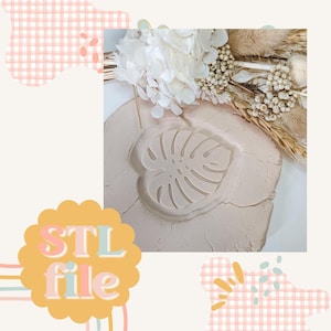 May include: A beige clay surface with a raised impression of a monstera leaf. The clay is textured and has a slightly rough surface. The leaf is detailed and has a realistic appearance. The image includes the text "STL file" in a colourful font.