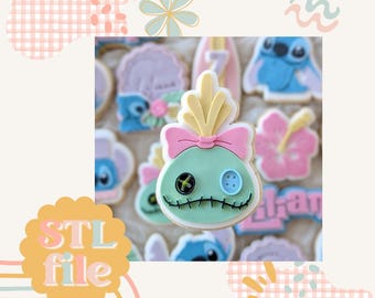 May include: A blue and green cookie decorated with a pink bow and a yellow tuft of hair. The cookie is shaped like a character with a stitched mouth and two buttons for eyes. The cookie is surrounded by other cookies, including a blue cookie with a flower design, a pink cookie with a flower design, and a blue cookie with a wave design.
