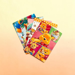 May include: A set of rectangular planner inserts featuring Winnie the Pooh illustrations. The cards display vibrant scenes of the yellow bear with honey pots labeled HUNNY, colorful balloons, and floral patterns against red, green, and white backgrounds.