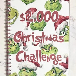 May include: A spiral-bound notebook with a white cover featuring a pattern of green Grinch faces wearing Santa hats. The text "$2,000 Christmas Challenge" is printed in burgundy.
