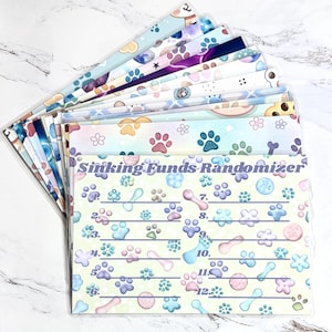 May include: A collection of clear plastic budget envelopes with colorful patterns. Designs include paw prints, bones, and cartoon characters. The text "Sinking Funds Randomizer" is printed on the front of the envelopes, along with numbers 1-12.