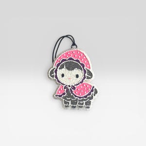 May include: A glittery, decorative sheep ornament with a pink polka-dot cape and hood. The sheep has a black body, gray face, and purple accents. A black string is attached for hanging. This whimsical ornament is perfect for a nursery or children's room.