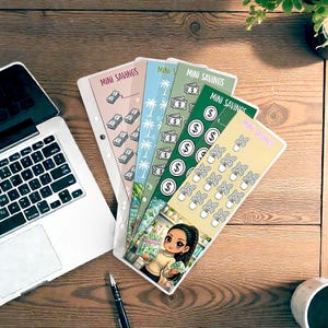 May include: A collection of colorful mini savings trackers with illustrations of money, palm trees, and plants. The trackers are arranged on a wooden surface with a laptop, pen, and coffee cup.