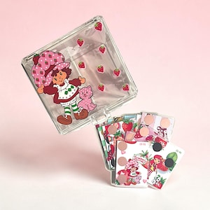 May include: A clear plastic case with a Strawberry Shortcake graphic and scattered strawberry stickers. The case is open, revealing a stack of cards with Strawberry Shortcake and strawberry designs. The cards have black magnetic dots.