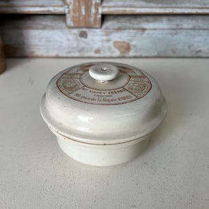 Puede incluir: Un recipiente de cerámica blanca con tapa. La tapa tiene una inscripción marrón que dice "Par VIANEY FRERES, Fabricants, 58, Rue de la Rapee, PARIS".