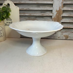 May include: A white ceramic cake stand with a scalloped edge. The stand features a wide, shallow bowl and a short, rounded base. The background includes a cutting board, a plant in a ceramic pot, and a wooden shutter.