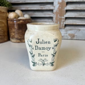 Puede incluir: Un tarro de cerámica blanco con diseños florales verdes y el texto "Julien Damoy - Paris".