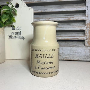 Puede incluir: Un tarro de mostaza de cerámica crema, de época, con la inscripción "MAILLE Moutarde à l'ancienne" en letras negras. El tarro tiene un borde decorativo en forma de cuerda y una parte superior ligeramente ensanchada. El fondo incluye una tabla de cortar blanca y una persiana de madera.