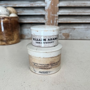 May include: Two vintage white ceramic jars with black lettering. The top jar reads "William Adams Shrewsbury". The bottom jar reads "James Woolley, Sons & Co. Pharmaceutical Chemists Manchester".