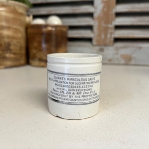 Könnte beinhalten: Ein kleines, antikes weißes Keramikgefäß mit einem schwarz-grauen Etikett. Das Etikett trägt die Aufschrift "Clarke's Miraculous Salve" und listet die Verwendungszwecke des Salbenprodukts auf. Das Gefäß steht auf einer weißen Oberfläche.