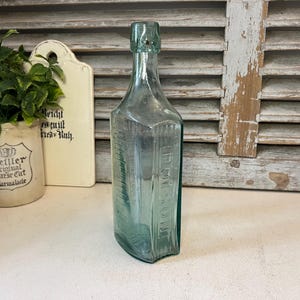 May include: A vintage light blue-green glass bottle with a rectangular base and a narrow neck. The word POISON is embossed in capital letters vertically along one side of the glass body. The bottle features a distinct rimmed top opening.