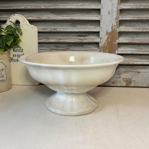 May include: A cream-coloured, pedestal bowl with a wide rim. The bowl has a slightly aged appearance, with subtle variations in colour. The base is a solid, rounded shape. The bowl is likely ceramic or porcelain.