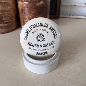 May include: A white ceramic jar with a lid. The lid has black lettering that reads "Creme D'Amandes Ameres Pour La Barbe Roger & Gallet 38 Rue D'Hauteville 38 Paris".