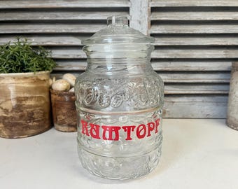 May include: A clear glass jar with a lid, featuring a decorative pattern and the word "RAUMTOPF" in red lettering. The jar is designed with a rounded shape and is likely used for storage or display.