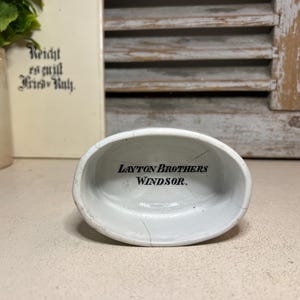 May include: A small, white, oval-shaped ceramic dish with the text "LAYTON BROTHERS WINDSOR" in black lettering inside. The dish has a slightly cracked appearance, suggesting age or wear. The background includes a blurred image of a book and a decorative shutter.