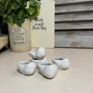 Peut inclure: Plusieurs petits porte-anneaux en céramique blanche. Les supports sont disposés sur une surface de couleur claire. En arrière-plan, une planche à découper de style vintage avec du texte allemand et un pot de marmelade avec une plante sont visibles.