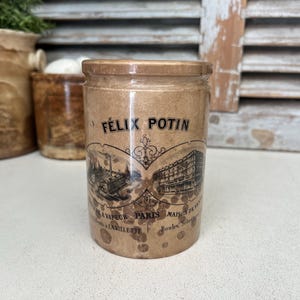 May include: A vintage, cylindrical ceramic jar with a lid. The jar is tan with a label featuring the text "Félix Potin" and illustrations. The label also includes text in French. The jar is likely for storage.