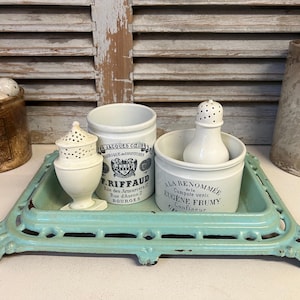 May include: A set of four vintage ceramic jars with lids, two with black and white labels, one with a black and white label and a crest, and one with a black and white label. The jars are sitting on a teal blue cast iron tray.