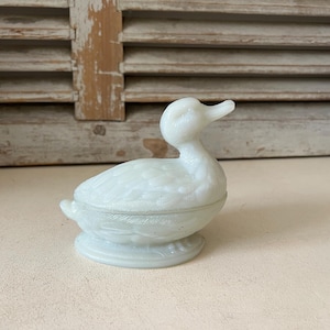 May include: A translucent white milk glass trinket dish shaped like a duck. The lidded container features moulded feather details on the body and wings, with the duck's head turned slightly to the right, resting on an oval base.