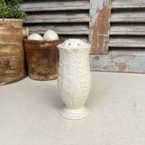 Puede incluir: Un salero de cerámica blanca con un diseño de hojas. El salero tiene una parte superior acampanada con varios agujeros y una base de pedestal. Otros elementos en el fondo incluyen una maceta de cerámica marrón con vegetación y un pequeño recipiente marrón con huevos blancos.