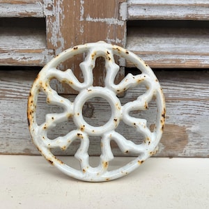 May include: A white, circular trivet with an ornate, openwork design. The trivet has a central circle with radiating, stylized floral or star-like elements. The edges show areas of rust, adding an aged, vintage aesthetic.