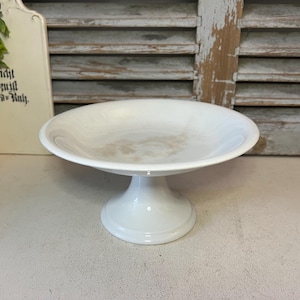 May include: A white ceramic cake stand with a wide, shallow dish and a pedestal base. The stand is clean and simple, perfect for displaying baked goods or other decorative items. The stand is approximately 20 cm in diameter.