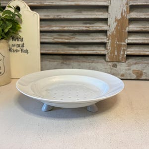 May include: A white ceramic plate with a perforated centre and three small feet. The plate is round with a slightly raised edge. The background includes a vintage-style cutting board and a decorative pot.