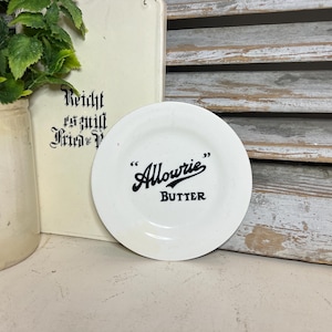 May include: A small, white plate with the word "Allowrie" in black script and the word "BUTTER" below it. The plate is set against a backdrop of a vintage-style book and a wooden shutter.