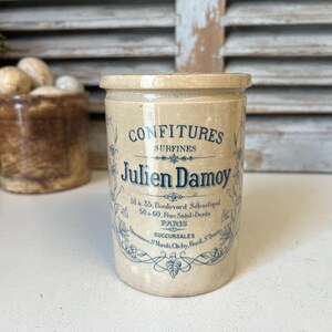 May include: A vintage beige ceramic jar with a lid, featuring blue text that reads "CONFITURES SURFINES Julien Damoy PARIS". The jar has a cylindrical shape and is likely used for preserving food items. Decorative floral elements surround the text.