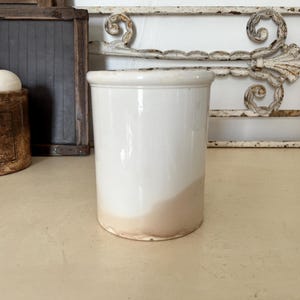 May include: A white ceramic crock with a slightly tapered shape and a rounded rim. The bottom portion of the crock has a light brown hue. Other items in the background include a vintage container and a decorative metal piece.