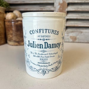 May include: White ceramic jar with blue and black lettering that reads "CONFITURES SURFINES Julien Damoy 31 à 35, Boulevard Sébastopol 50 à 60, Rue Saint-Denis PARIS SUCCURSALES SMdi Clichy Rueil SD".