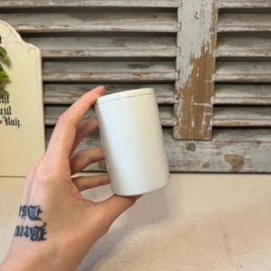 May include: A small, cylindrical, white ceramic container with a smooth, solid finish. The object features a simple, minimalist design with a flat top and straight sides, presented as a plain, unadorned vessel suitable for storage or display purposes.