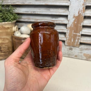 Puede incluir: Un pequeño tarro de cerámica marrón con tapa. El tarro tiene una etiqueta que dice "W. & J. AUCHMUTY, WESTMINSTER".