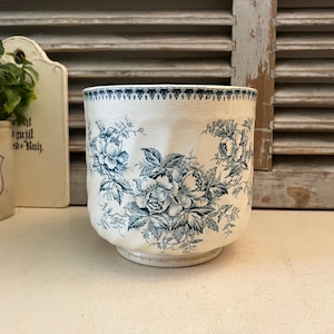 Puede incluir: Un jarrón de cerámica blanco con un estampado floral azul. El jarrón tiene forma redondeada y un borde ligeramente acampanado. El diseño floral presenta rosas y hojas. El jarrón está sobre una superficie blanca.