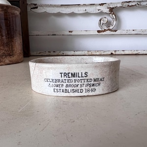 May include: An antique, off-white ceramic dish with the text "TREMILLS CELEBRATED POTTED MEAT 5, LOWER BROOK ST IPSWICH ESTABLISHED 1849" in black lettering. The dish has a cracked, aged appearance, suggesting its vintage. A second, similar jar is visible in the background.