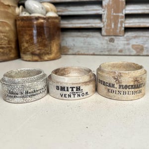 May include: Three vintage ceramic containers with lids. The containers are white with black lettering. The text on the containers reads "Allan & Hanburys Ltd. 77, Oxford Street, Cavendish Square, London", "Smith, Chemist, Ventnor", and "Duncan Flockhart & Co. Edinburgh".