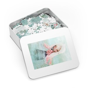 May include: A white metal tin with a lid that opens to reveal a completed jigsaw puzzle. The puzzle depicts a girl with long pink hair holding a small pink pig. The girl is wearing a black dress and is standing in a blue and white water scene.