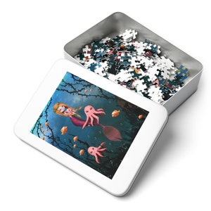 May include: A white metal tin with a blue and pink illustration of a mermaid riding an octopus with two smaller octopuses in the background. The tin is open and filled with a jigsaw puzzle.