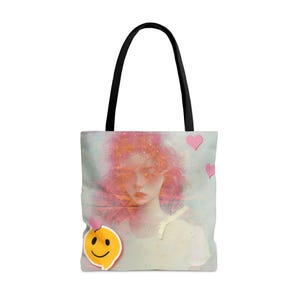 May include: A tote bag with a pink and white watercolor design of a woman's face. The design includes a yellow smiley face and pink hearts.