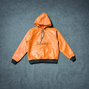 May include: Orange leather hoodie with a drawstring and a black trim on the cuffs and hem. The interior of the hood is a contrasting red color. The hoodie is made of a smooth, shiny material.