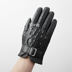 Op de afbeelding: Zwarte leren rijhandschoenen met een krokodillenleertextuur. De handschoenen hebben een riem met een zilveren gesp bij de pols en kleine perforaties op de vingers. Het woord "König" is zichtbaar.