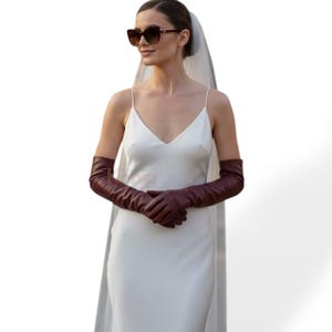 Puede incluir: Una mujer con un vestido blanco de tirantes finos y largos guantes de cuero burdeos oscuro. Lleva gafas de sol y un velo. Los guantes se extienden más allá de los codos. El vestido tiene un escote en V.