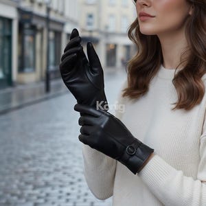 Könnte beinhalten: Schwarze Lederhandschuhe mit Schnallendetail am Handgelenk. Die Handschuhe sind glatt und scheinen aus hochwertigem Leder gefertigt zu sein. Die Handschuhe werden von einer Person getragen, die einen weißen Pullover trägt.