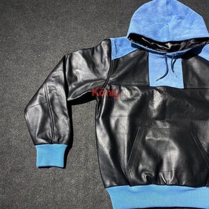 May include: Black leather hoodie with a blue hood and cuffs. The hoodie features a kangaroo pocket and a contrasting blue panel on the upper chest. The cuffs and hood lining are also blue.