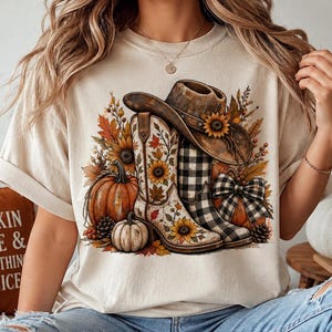 Puede incluir: Una camiseta color crema con un estampado gráfico de un sombrero de vaquero marrón sobre dos botas vaqueras. Una bota tiene un patrón de girasoles y la otra un patrón de cuadros blanco y negro. Calabazas, hojas de otoño y girasoles las rodean.