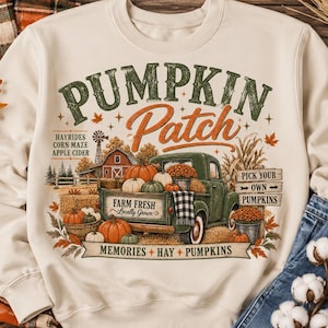 May include: A cream-colored crewneck sweatshirt featuring a graphic print of a green vintage truck loaded with pumpkins. The text reads PUMPKIN PATCH, HAYRIDES, CORN MAZE, APPLE CIDER, FARM FRESH, PICK YOUR OWN PUMPKINS, and MEMORIES HAY PUMPKINS.