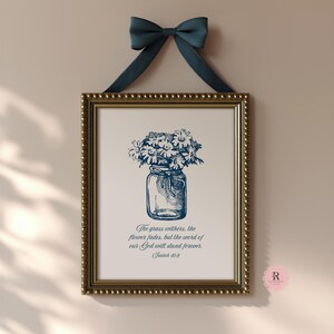 Puede incluir: Impresión enmarcada con un marco dorado con cuentas, que representa una ilustración azul y blanca de margaritas en un tarro de cristal. La impresión está suspendida por una cinta azul marino. Texto: "La hierba se seca, la flor se marchita, pero la palabra de nuestro Dios permanece para siempre. Isaías 40:8."