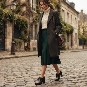 May include: A dark gray wool coat worn over a cream-colored cable-knit sweater and a dark green pleated midi skirt. The outfit is paired with black leather lace-up shoes featuring a light-colored panel and a brown leather shoulder bag.
